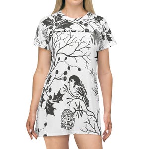 Könnte beinhalten: Ein weißes T-Shirt-Kleid mit einem schwarzen botanischen Druck aus Stechpalmenblättern, Beeren, Tannenzapfen und einem kleinen Vogel auf einem Ast. Der Text "grounded but restless" ist in Schwarz im oberen Brustbereich aufgedruckt.