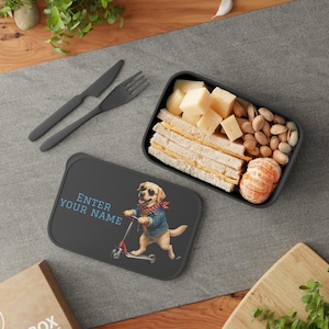 Könnte beinhalten: Eine rechteckige schwarze Lunchbox mit einem Deckel, der eine Grafik eines Golden Retrievers mit Sonnenbrille, Jeansjacke und rotem Halstuch auf einem Roller zeigt, mit dem Text ENTER YOUR NAME. Enthält ein schwarzes Messer und eine Gabel.