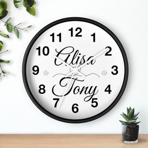 Könnte beinhalten: Eine runde Wanduhr mit schwarzem Rahmen und weißem Zifferblatt mit schwarzen Zahlen von 1 bis 12. In der Mitte stehen die Namen Alisa und Tony in schwarzer Schreibschrift, getrennt durch eine Zierlinie und zwei kleine Blumensymbole.