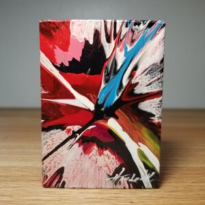 May include: A rectangular wooden block featuring an abstract acrylic pour painting with vibrant streaks of red, black, white, and bright blue. The paint design radiates from the center in dynamic, fluid lines, with a signature visible in the bottom corner.