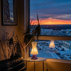May include: A modern table lamp with a white shade and purple base sits on a glass table. The lamp is lit, casting a warm glow. A vibrant sunset is visible through the window, with a cityscape in the background. A framed art piece is on the wall.
