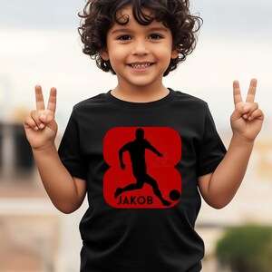 Pode incluir: Camiseta preta com um gr&aacute;fico vermelho de um jogador de futebol chutando uma bola, com o nome "JAKOB" abaixo. O gr&aacute;fico est&aacute; dentro de um oito vermelho. A camiseta &eacute; usada por uma crian&ccedil;a sorrindo.