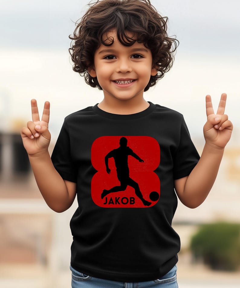 Pode incluir: Camiseta preta com um gr&aacute;fico vermelho de um jogador de futebol chutando uma bola, com o nome "JAKOB" abaixo. O gr&aacute;fico est&aacute; dentro de um oito vermelho. A camiseta &eacute; usada por uma crian&ccedil;a sorrindo.