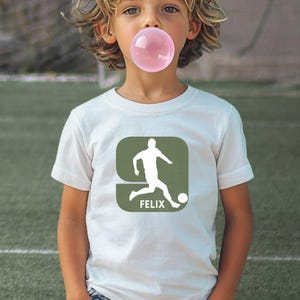 Puede incluir: Una camiseta blanca de manga corta con un gr&aacute;fico cuadrado de color verde oliva centrado. El gr&aacute;fico muestra una silueta blanca de un jugador de f&uacute;tbol pateando un bal&oacute;n y el nombre FELIX impreso en letras may&uacute;sculas blancas debajo.