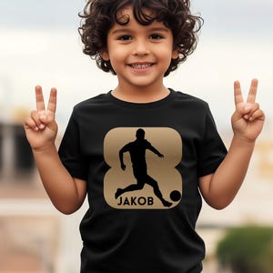 Pode incluir: Camiseta preta com um gr&aacute;fico dourado de um jogador de futebol chutando uma bola, com o nome "JAKOB" abaixo. O design est&aacute; dentro de uma forma dourada. A camiseta &eacute; usada por uma crian&ccedil;a.