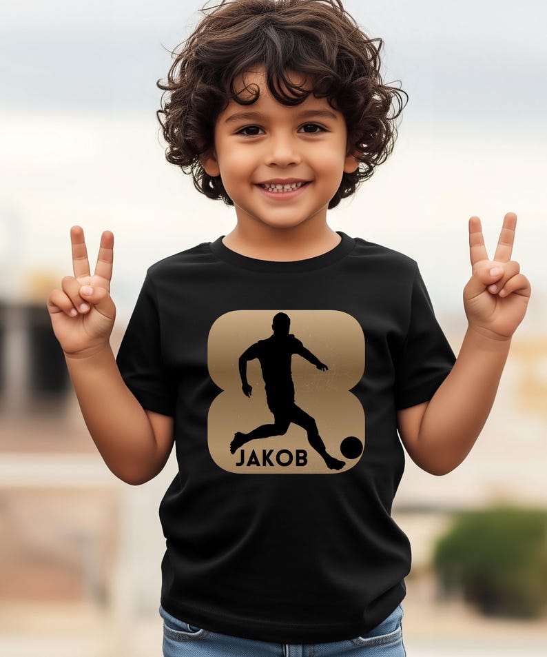 Pode incluir: Camiseta preta com um gr&aacute;fico dourado de um jogador de futebol chutando uma bola, com o nome "JAKOB" abaixo. O design est&aacute; dentro de uma forma dourada. A camiseta &eacute; usada por uma crian&ccedil;a.