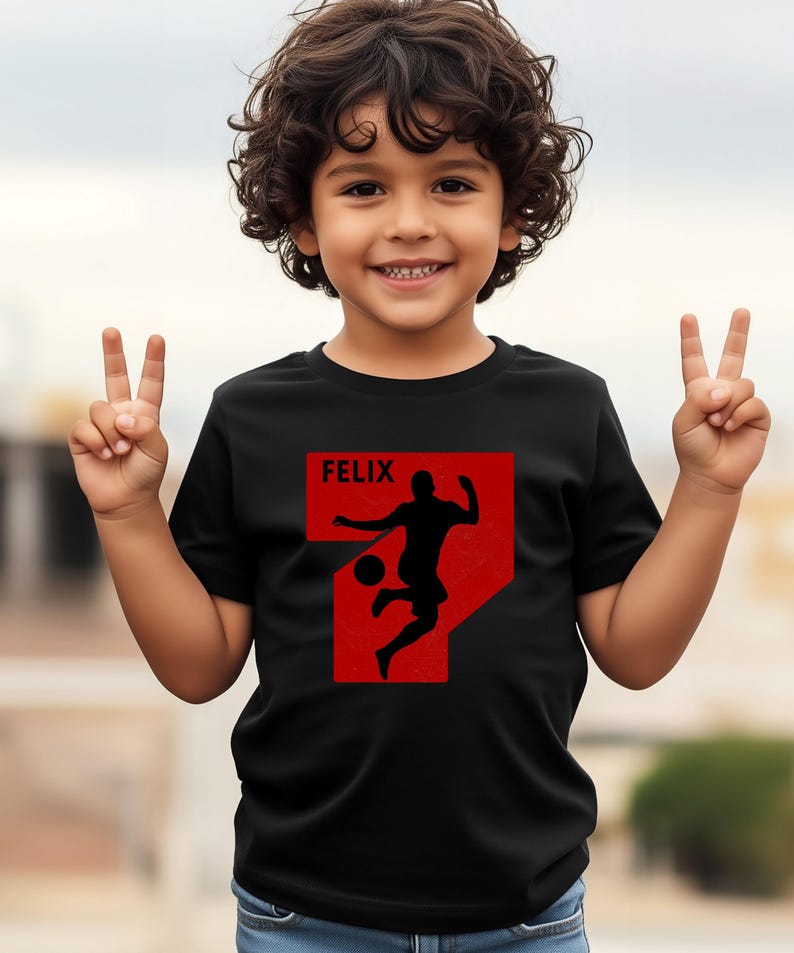 Pode incluir: Camiseta preta com um gr&aacute;fico vermelho de um jogador de futebol chutando uma bola, dentro de um n&uacute;mero 7 vermelho. O nome "FELIX" est&aacute; acima do gr&aacute;fico. A crian&ccedil;a est&aacute; sorrindo e levantando dois dedos em cada m&atilde;o.