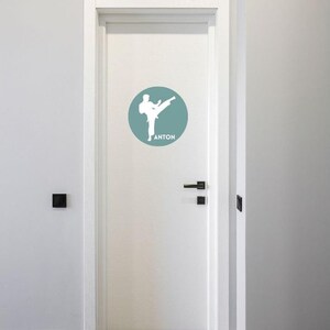 May include: A white door featuring a circular decal in a muted blue-green shade. The decal displays a white silhouette of a person performing a martial arts kick, with the name ANTON printed in white capital letters beneath the figure.