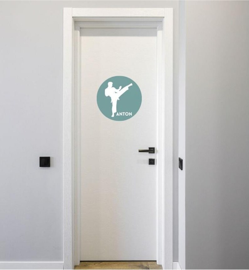 May include: A white door featuring a circular decal in a muted blue-green shade. The decal displays a white silhouette of a person performing a martial arts kick, with the name ANTON printed in white capital letters beneath the figure.