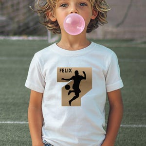 Puede incluir: Una camiseta blanca de manga corta con un estampado gr&aacute;fico de la silueta negra de un jugador de f&uacute;tbol pateando un bal&oacute;n. Sobre el jugador, el nombre FELIX est&aacute; impreso en letras may&uacute;sculas negras sobre un fondo rectangular de color tostado.