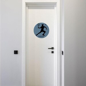 May include: A white door featuring a circular wall decal. The decal is slate blue and displays a black silhouette of a person playing tennis with a racket in hand. The name JAKOB is printed in black capital letters at the bottom of the circle.