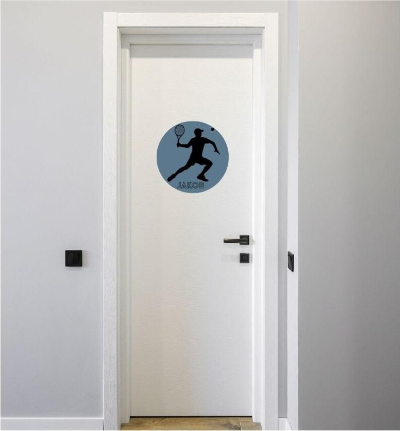 May include: A white door featuring a circular wall decal. The decal is slate blue and displays a black silhouette of a person playing tennis with a racket in hand. The name JAKOB is printed in black capital letters at the bottom of the circle.