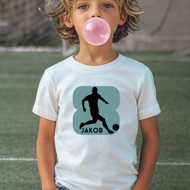 Puede incluir: Una camiseta blanca de manga corta con un gr&aacute;fico de cuadrado redondeado de color azul claro en el pecho. El gr&aacute;fico muestra la silueta negra de un jugador de f&uacute;tbol pateando un bal&oacute;n con el nombre JAKOB impreso en letras negras may&uacute;sculas.