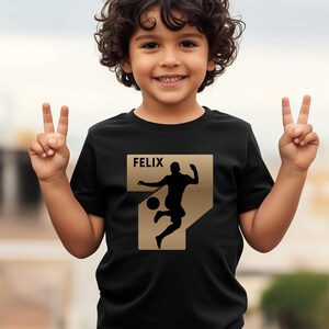 Pode incluir: Camiseta preta com um design dourado do n&uacute;mero sete com a silhueta de um jogador de futebol. O nome "FELIX" &eacute; impresso acima do gr&aacute;fico. A crian&ccedil;a est&aacute; sorrindo e levantando dois dedos de cada m&atilde;o.