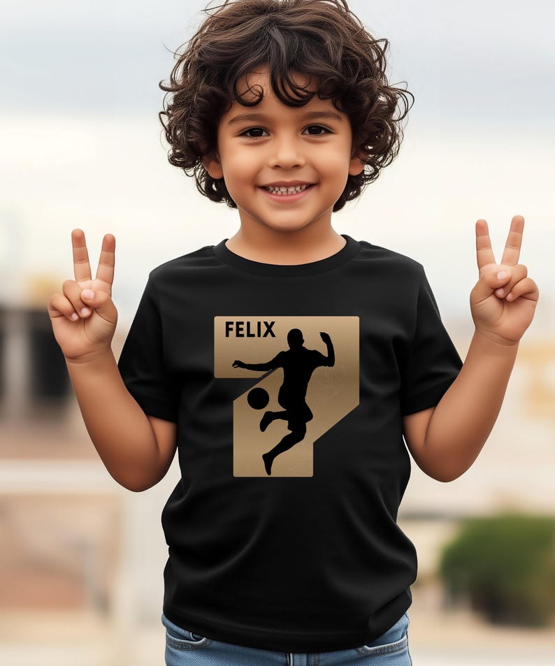 Pode incluir: Camiseta preta com um design dourado do n&uacute;mero sete com a silhueta de um jogador de futebol. O nome "FELIX" &eacute; impresso acima do gr&aacute;fico. A crian&ccedil;a est&aacute; sorrindo e levantando dois dedos de cada m&atilde;o.