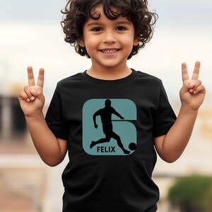 Pode incluir: Camiseta preta com um gr&aacute;fico turquesa de um jogador de futebol chutando uma bola, com o nome "FELIX" abaixo. O desenho est&aacute; dentro de uma forma turquesa. A camiseta &eacute; usada por uma crian&ccedil;a sorrindo.