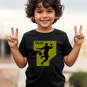 Pode incluir: Camiseta preta com uma silhueta de jogador de futebol em um design amarelo e verde. O nome "FELIX" &eacute; impresso acima da silhueta. A crian&ccedil;a est&aacute; sorrindo e levantando dois dedos.