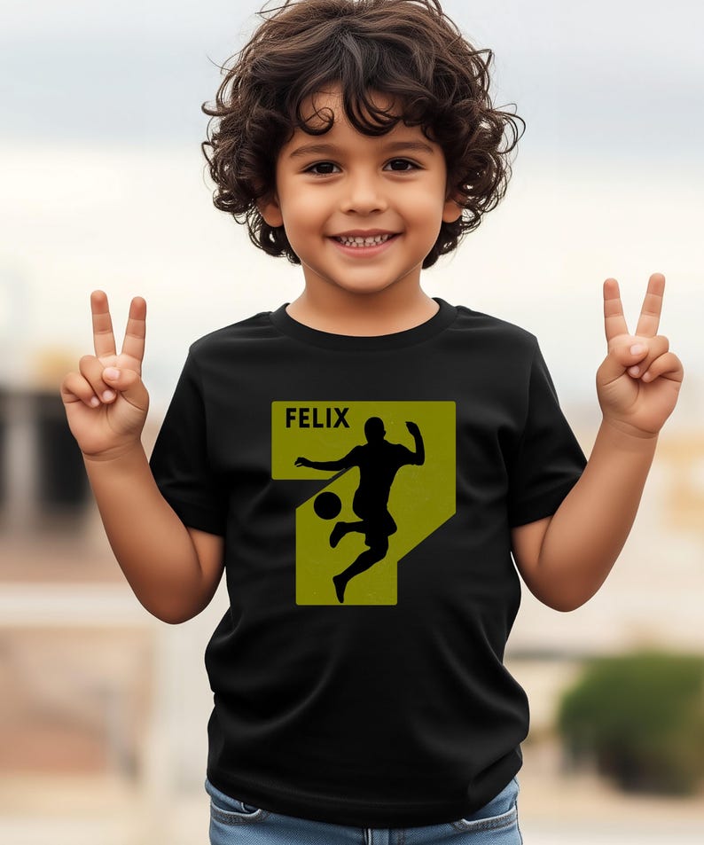 Pode incluir: Camiseta preta com uma silhueta de jogador de futebol em um design amarelo e verde. O nome "FELIX" &eacute; impresso acima da silhueta. A crian&ccedil;a est&aacute; sorrindo e levantando dois dedos.