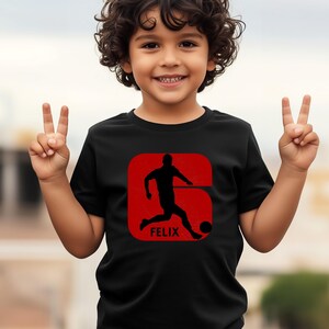 Pode incluir: Camiseta preta com um gr&aacute;fico vermelho de um jogador de futebol chutando uma bola, com o nome "FELIX" abaixo. O design est&aacute; dentro de um ret&acirc;ngulo vermelho arredondado. A crian&ccedil;a est&aacute; sorrindo e fazendo o sinal de paz.