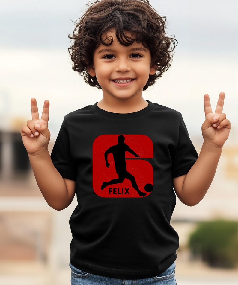 Pode incluir: Camiseta preta com um gr&aacute;fico vermelho de um jogador de futebol chutando uma bola, com o nome "FELIX" abaixo. O design est&aacute; dentro de um ret&acirc;ngulo vermelho arredondado. A crian&ccedil;a est&aacute; sorrindo e fazendo o sinal de paz.