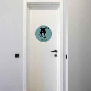 May include: A white door featuring a circular decal with a muted teal background. The decal displays a black silhouette of a person performing a skateboard trick, with the name JULIAN printed in black capital letters underneath the figure.