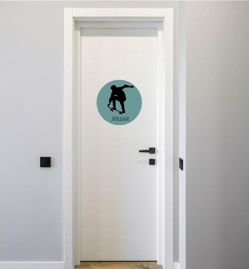 May include: A white door featuring a circular decal with a muted teal background. The decal displays a black silhouette of a person performing a skateboard trick, with the name JULIAN printed in black capital letters underneath the figure.