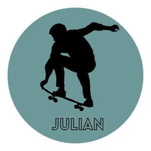May include: A circular graphic featuring a muted teal background with a black silhouette of a person performing a skateboard trick. The person is crouched over the board with one hand reaching down. The name JULIAN is printed in black below.