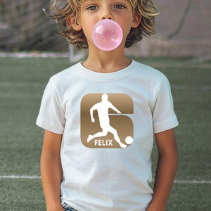 Puede incluir: Una camiseta blanca de manga corta con un gr&aacute;fico cuadrado marr&oacute;n de esquinas redondeadas. El gr&aacute;fico muestra la silueta blanca de un jugador de f&uacute;tbol pateando un bal&oacute;n y el nombre FELIX impreso en letras may&uacute;sculas blancas debajo.