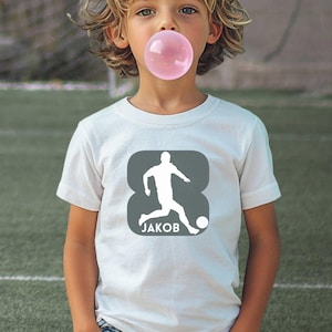 Puede incluir: Una camiseta blanca de manga corta con un gr&aacute;fico cuadrado de color gris oscuro y esquinas redondeadas. El gr&aacute;fico muestra la silueta blanca de un jugador de f&uacute;tbol pateando un bal&oacute;n, con el nombre JAKOB impreso en letras may&uacute;sculas blancas.
