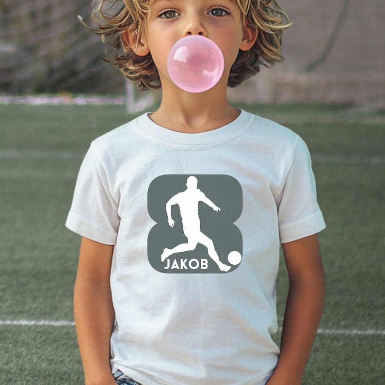 Puede incluir: Una camiseta blanca de manga corta con un gr&aacute;fico cuadrado de color gris oscuro y esquinas redondeadas. El gr&aacute;fico muestra la silueta blanca de un jugador de f&uacute;tbol pateando un bal&oacute;n, con el nombre JAKOB impreso en letras may&uacute;sculas blancas.