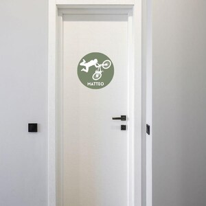 May include: A circular sage green wall decal applied to a white door, featuring a white silhouette of a person performing a bicycle stunt in mid-air. Below the graphic, the name MATTEO is printed in white capital letters in a sans-serif font.