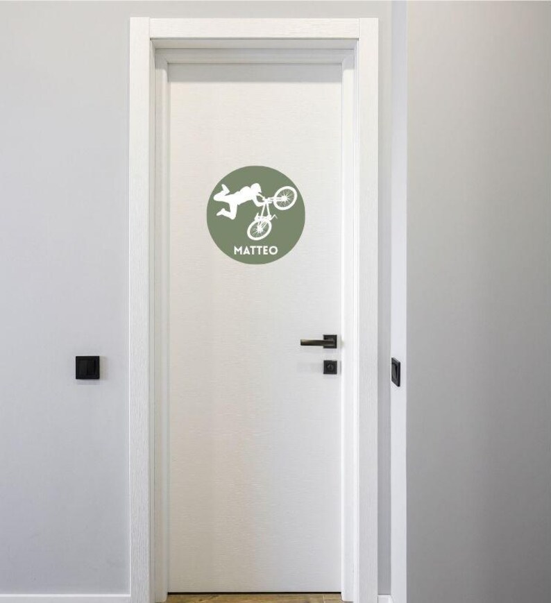 May include: A circular sage green wall decal applied to a white door, featuring a white silhouette of a person performing a bicycle stunt in mid-air. Below the graphic, the name MATTEO is printed in white capital letters in a sans-serif font.