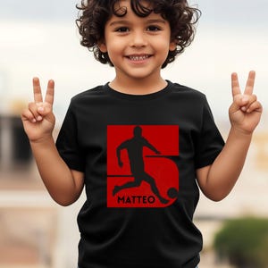 Pode incluir: Camiseta preta com um gr&aacute;fico vermelho de um jogador de futebol chutando uma bola, com o nome "MATTEO" abaixo. O n&uacute;mero "5" est&aacute; incorporado no design. A crian&ccedil;a est&aacute; sorrindo e fazendo um sinal de paz.