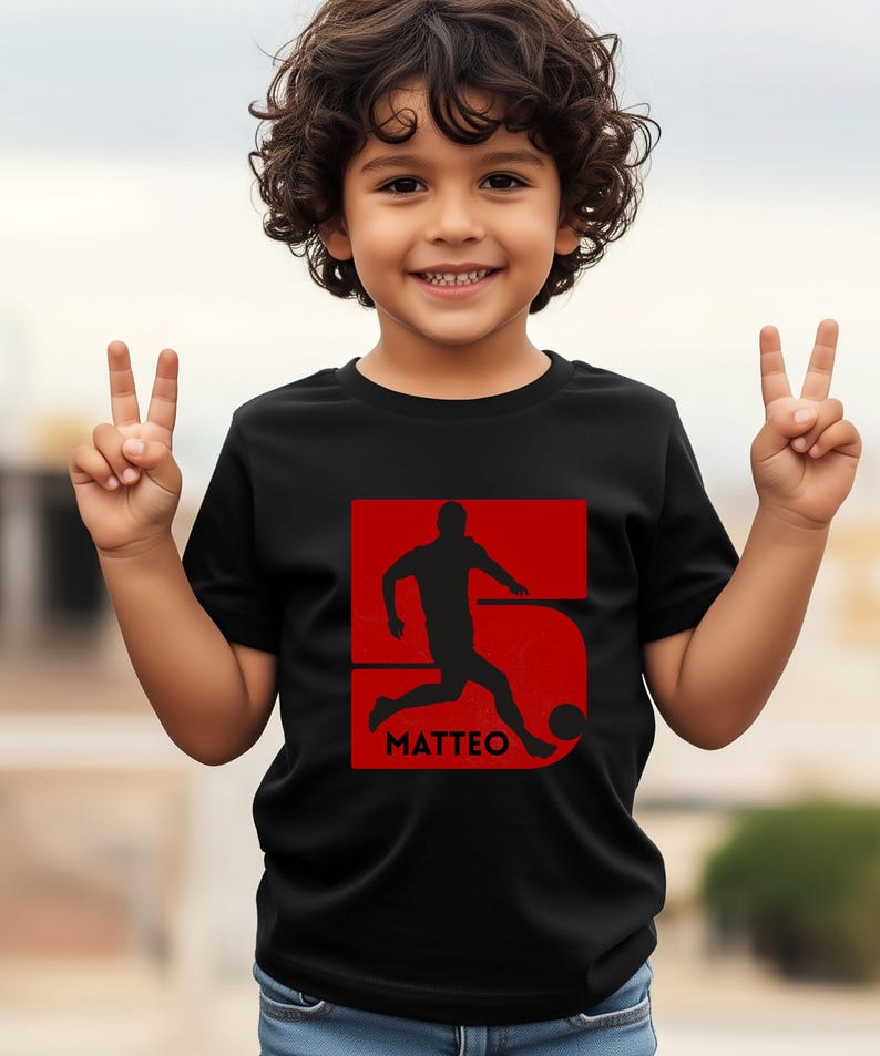 Pode incluir: Camiseta preta com um gr&aacute;fico vermelho de um jogador de futebol chutando uma bola, com o nome "MATTEO" abaixo. O n&uacute;mero "5" est&aacute; incorporado no design. A crian&ccedil;a est&aacute; sorrindo e fazendo um sinal de paz.