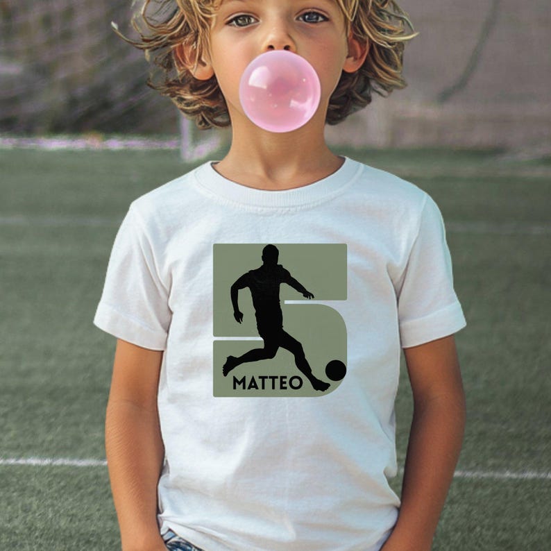 Puede incluir: Una camiseta blanca de manga corta con un dise&ntilde;o gr&aacute;fico de un n&uacute;mero cinco grande en color verde oliva. Dentro del n&uacute;mero hay una silueta negra de un jugador de f&uacute;tbol pateando un bal&oacute;n, con el nombre MATTEO impreso en letras negras.
