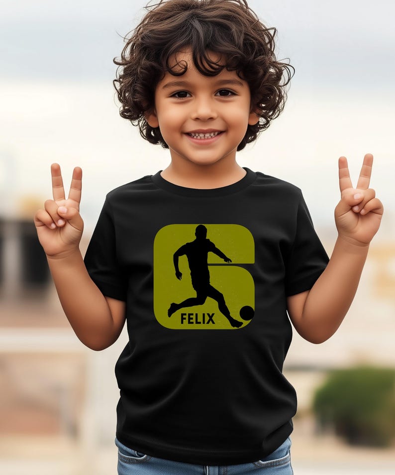 Pode incluir: Camiseta preta com um design de jogador de futebol em silhueta dentro de um quadrado verde e o nome "FELIX" abaixo. Uma crian&ccedil;a sorri e levanta dois dedos de cada m&atilde;o.