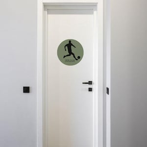 May include: A white door featuring a circular sage green decal. The decal displays a black silhouette of a football player kicking a ball, with the name JULIAN printed in black capital letters directly underneath the player figure.