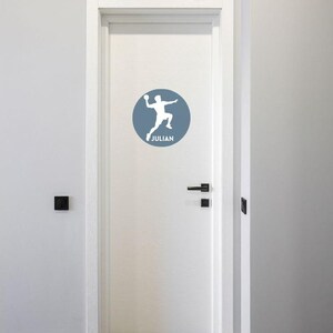 May include: A white door featuring a circular blue decal. The decal displays a white silhouette of a person jumping while holding a ball, with the name JULIAN printed in white capital letters underneath the figure within the blue circle.