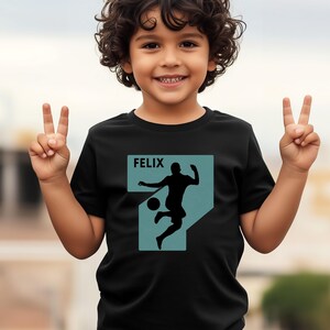 Pode incluir: T-shirt preto com um gr&aacute;fico turquesa de um jogador de futebol a chutar uma bola, com o nome "FELIX" acima. O gr&aacute;fico tem a forma do n&uacute;mero 7. A t-shirt &eacute; usada por uma crian&ccedil;a pequena.