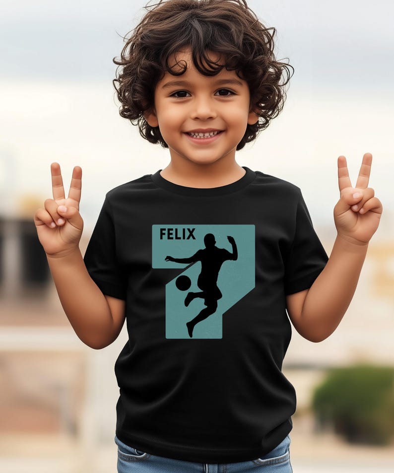 Pode incluir: T-shirt preto com um gr&aacute;fico turquesa de um jogador de futebol a chutar uma bola, com o nome "FELIX" acima. O gr&aacute;fico tem a forma do n&uacute;mero 7. A t-shirt &eacute; usada por uma crian&ccedil;a pequena.