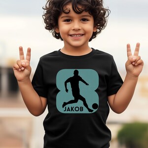Pode incluir: Camiseta preta com um gr&aacute;fico turquesa de um jogador de futebol chutando uma bola, com o nome "JAKOB" abaixo. O n&uacute;mero "8" est&aacute; integrado no design. A crian&ccedil;a est&aacute; sorrindo e fazendo o sinal de vit&oacute;ria.