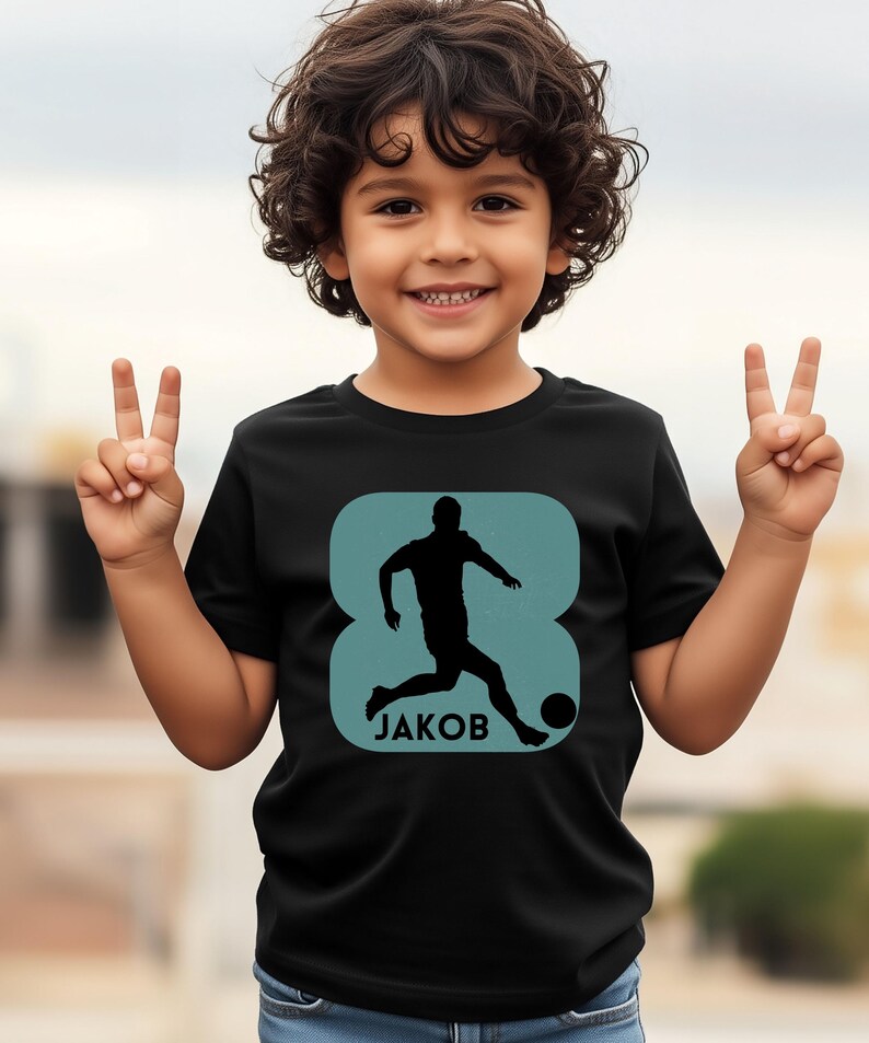 Pode incluir: Camiseta preta com um gr&aacute;fico turquesa de um jogador de futebol chutando uma bola, com o nome "JAKOB" abaixo. O n&uacute;mero "8" est&aacute; integrado no design. A crian&ccedil;a est&aacute; sorrindo e fazendo o sinal de vit&oacute;ria.