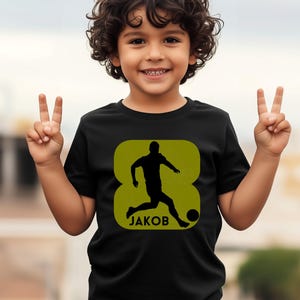 Pode incluir: Camiseta preta com um design verde em forma de oito que mostra a silhueta de um jogador de futebol. O nome "JAKOB" est&aacute; impresso abaixo. A camiseta &eacute; usada por uma crian&ccedil;a.