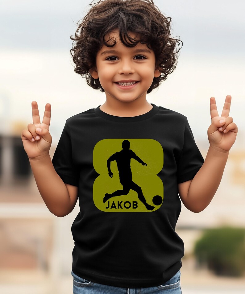 Pode incluir: Camiseta preta com um design verde em forma de oito que mostra a silhueta de um jogador de futebol. O nome "JAKOB" est&aacute; impresso abaixo. A camiseta &eacute; usada por uma crian&ccedil;a.