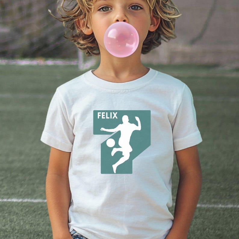 Puede incluir: Una camiseta blanca de manga corta con un gr&aacute;fico rectangular de color verde apagado en el pecho. El gr&aacute;fico muestra la silueta blanca de un jugador de f&uacute;tbol pateando un bal&oacute;n y el nombre FELIX impreso en letras may&uacute;sculas blancas.