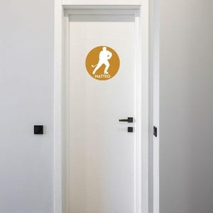 May include: A white door featuring a circular mustard yellow decal. The decal displays a white silhouette of a hockey player in motion, holding a hockey stick, with the name MATTEO printed in white capital letters underneath the figure.