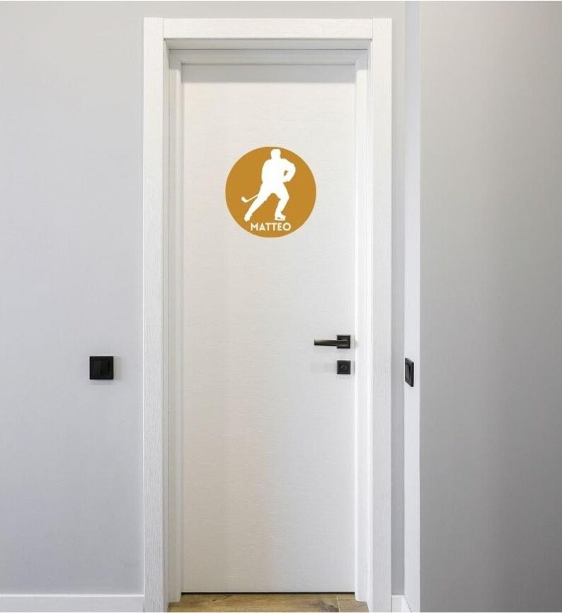May include: A white door featuring a circular mustard yellow decal. The decal displays a white silhouette of a hockey player in motion, holding a hockey stick, with the name MATTEO printed in white capital letters underneath the figure.