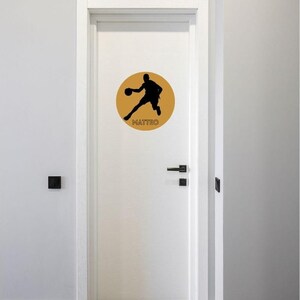 May include: A white door featuring a circular gold decal with a black silhouette of a basketball player in motion. The name MATTEO is printed in black capital letters at the bottom of the gold circle, centred below the basketball player figure.