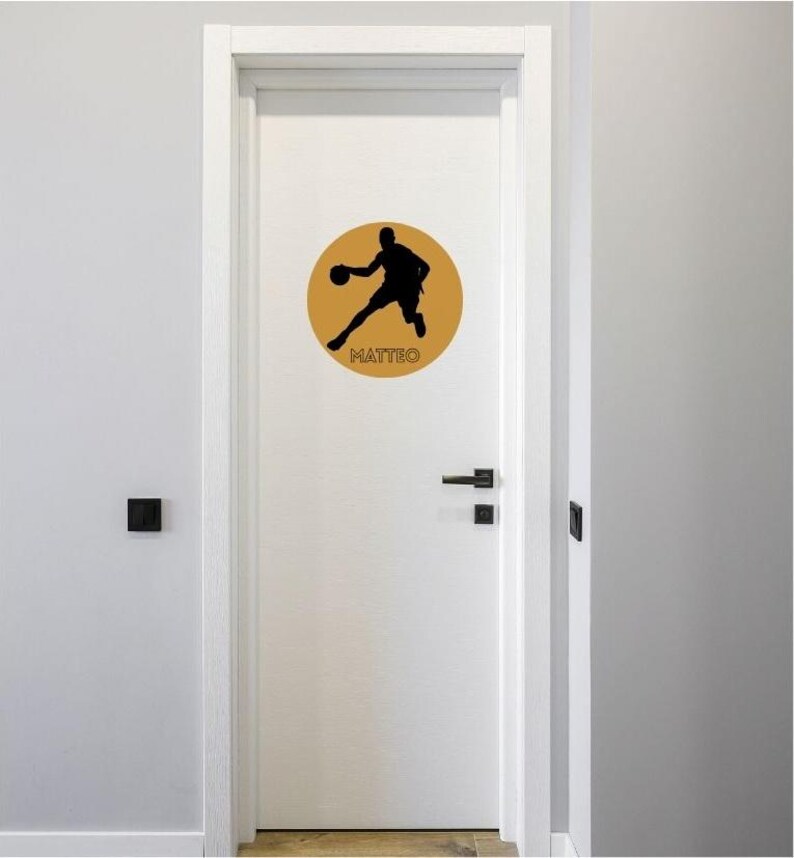May include: A white door featuring a circular gold decal with a black silhouette of a basketball player in motion. The name MATTEO is printed in black capital letters at the bottom of the gold circle, centred below the basketball player figure.
