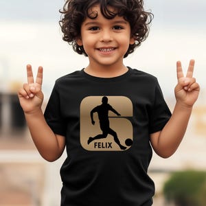 Pode incluir: Camiseta preta com um desenho dourado de um jogador de futebol chutando uma bola, com o nome "FELIX" abaixo. O desenho est&aacute; dentro de uma forma dourada. A camiseta &eacute; usada por uma crian&ccedil;a.